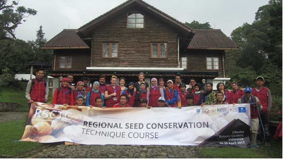 A row of people all stood smiling in front of a wooden building holding a banner reading Regional Seed Conservation Technique Course